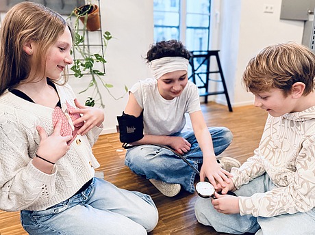 Drei Kinder sitzen auf einem Holzboden. Ein Mädchen trägt einen Verband und hat eine Blutdruckmanschette am Arm, während ein Junge auf das Manometer schaut. Ein weiteres Mädchen lächelt und deutet auf ihre Brust. Es sieht so aus, als würden sie zusammen spielen oder lernen.