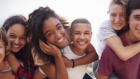 Kindergesundheitsbericht 2023 Sechs lächelnde Teenager posieren eng beieinander im Freien. Zwei tragen die anderen auf dem Rücken, und alle sehen in ihrer Freizeitkleidung glücklich und fröhlich aus, während im Hintergrund die Sonne scheint.