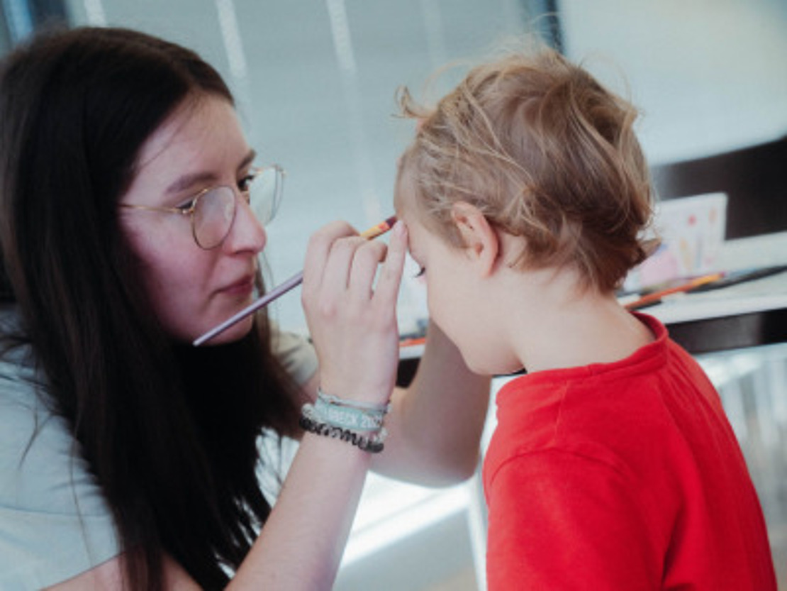 Eine Frau mit Brille bemalt das Gesicht eines Kindes, während dieses in einem roten Hemd und einer grauen Hose auf einem Stuhl sitzt. Sie befinden sich in einem Raum, und die Frau hält in der anderen Hand eine Karte.