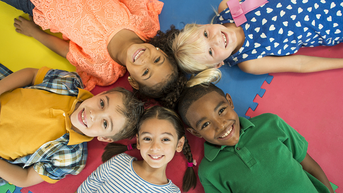 Fünf lächelnde Kinder liegen im Kreis auf einer bunten Bodenmatte und blicken in die Kamera.