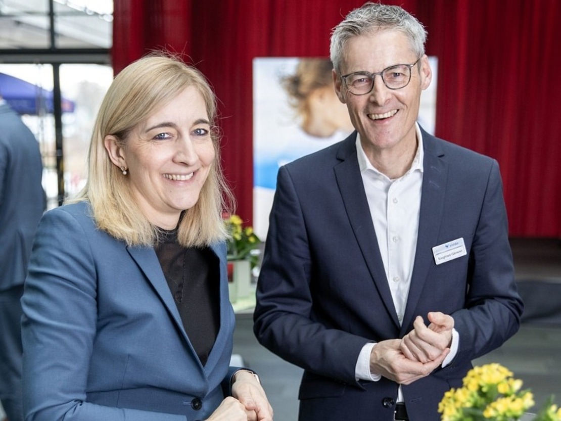 Zwei Personen in Geschäftskleidung lächeln in die Kamera, während sie an einem Tisch mit Blumen und Wassergläsern stehen. Im Hintergrund sind eine verschwommene Gestalt und rote Vorhänge zu sehen.