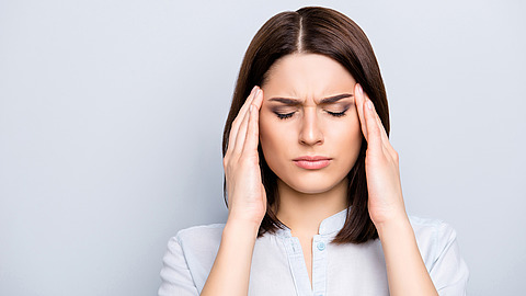 Eine Frau mit glattem braunem Haar, die sich die Schläfen hält und die Augen schließt; sie wirkt, als hätte sie Schmerzen oder stünde unter Stress, vor einem schlichten hellen Hintergrund.