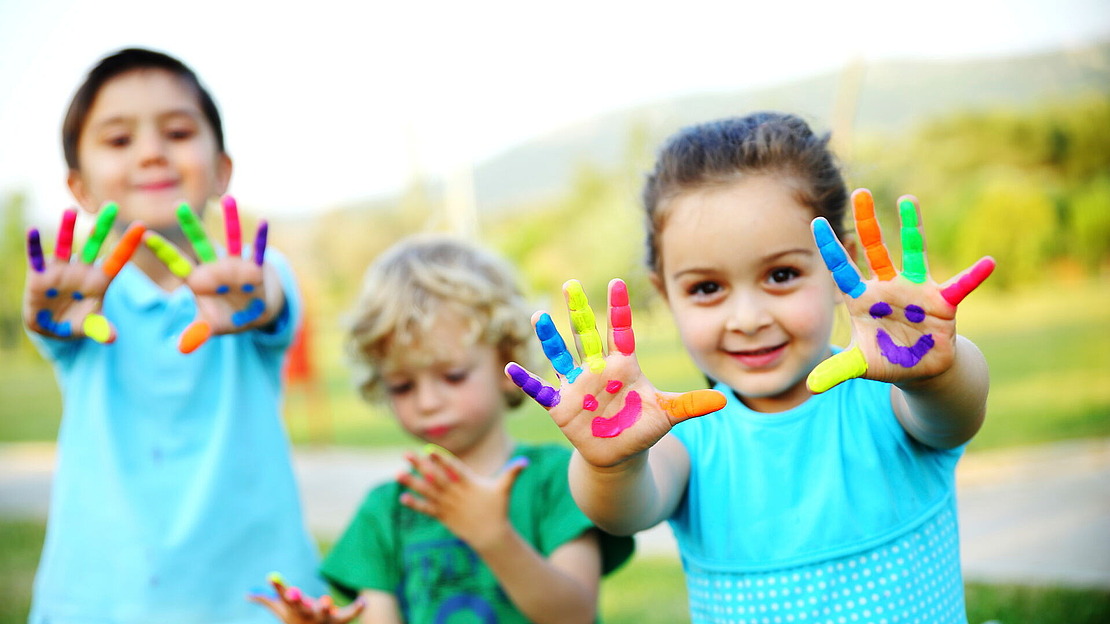 Drei kleine Kinder im Freien, die lächeln und ihre Hände hochhalten, die mit leuchtenden Farben und Smiley-Gesichtern bemalt sind. Zwei Kinder blicken in die Kamera, während eines auf seine Hände schaut. Im Hintergrund sieht man eine Wiese mit Bäumen und Hügeln.