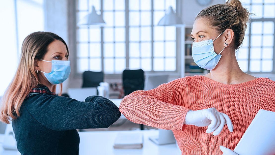 Zwei Frauen mit Gesichtsmasken und Handschuhen begrüßen sich in einem hellen Büroumfeld mit einer Berührung der Ellbogen und halten sich dabei an die Richtlinien zur sozialen Distanzierung.