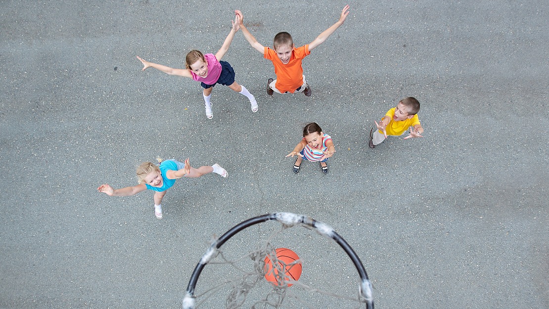 Fünf Kinder strecken die Hände aus, als sich ein Basketball dem Korb nähert – aus der Vogelperspektive auf einem Außenplatz betrachtet.