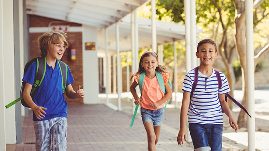 Drei Kinder mit Rucksäcken, die an einem sonnigen Tag vor der Schule lächelnd einen überdachten Gang entlanglaufen.