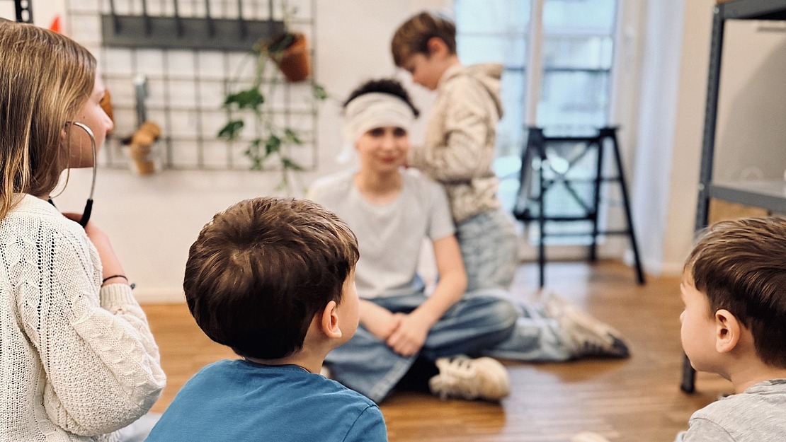 Fünf Kinder sitzen auf einem Holzboden in einem hellen Raum und unterhalten sich. An der Wand hinter ihnen hängen Pflanzen. Einige Kinder sitzen mit dem Rücken zur Kamera, während andere in ein Gespräch vertieft sind.