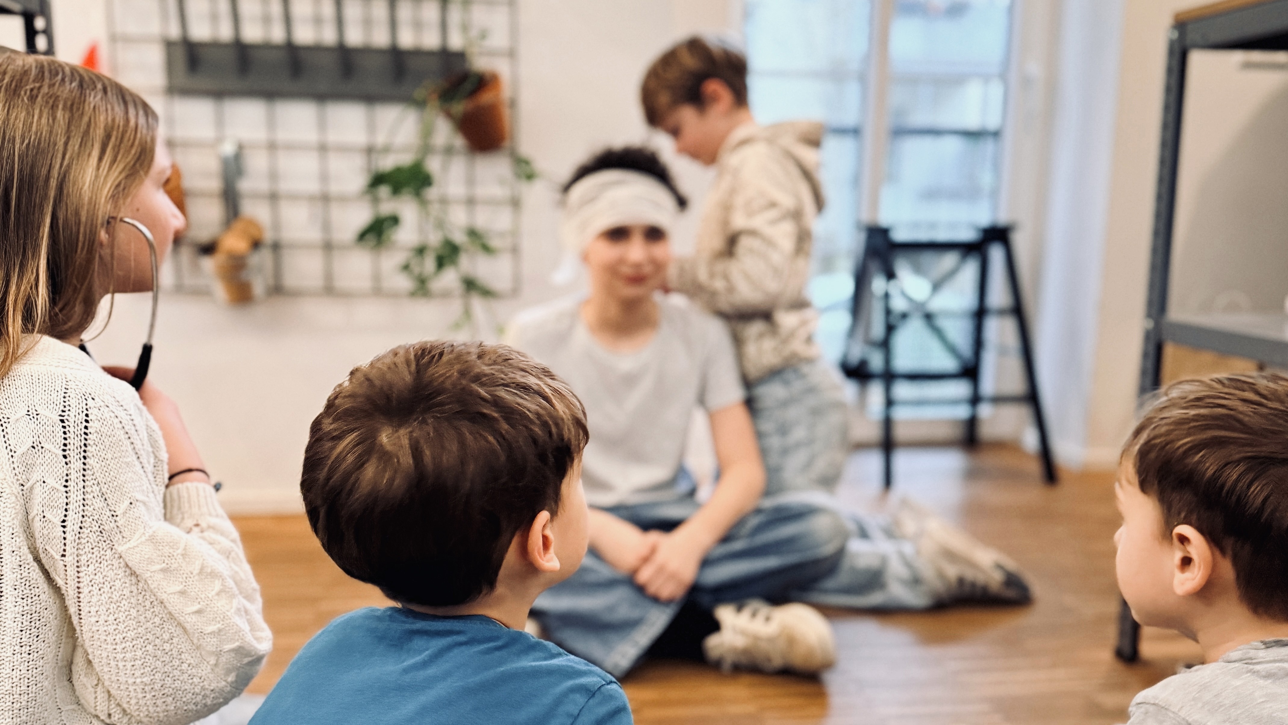 Fünf Kinder sitzen auf einem Holzboden in einem hellen Raum und unterhalten sich. An der Wand hinter ihnen hängen Pflanzen. Einige Kinder sitzen mit dem Rücken zur Kamera, während andere in ein Gespräch vertieft sind.