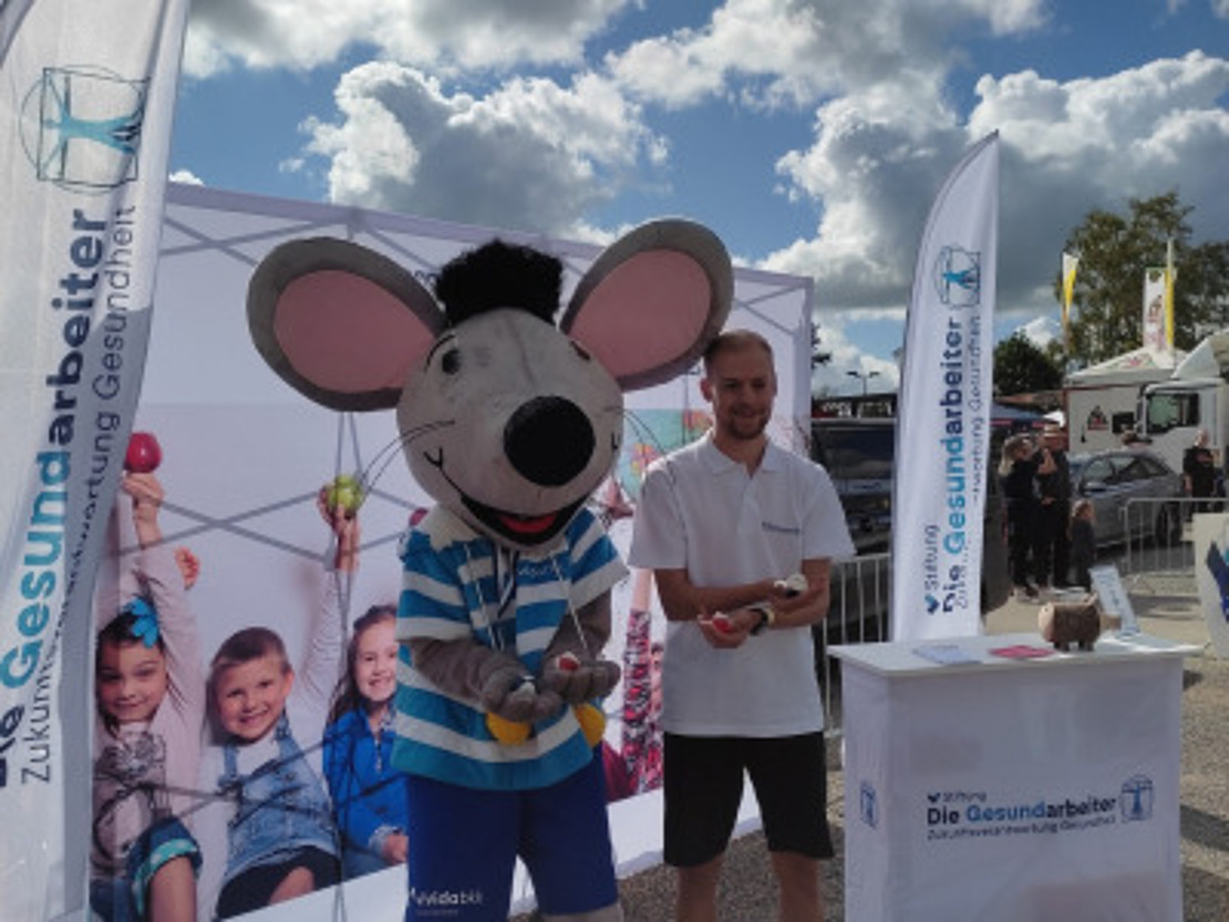 Eine Person steht neben einer großen Maus-Maskottchenfigur an einem Stand im Freien, an dem Banner und eine Kulisse mit Kindern zu sehen sind. Die Szene ist hell, am Himmel sind Wolken zu sehen, und im Vordergrund steht ein Werbetisch.