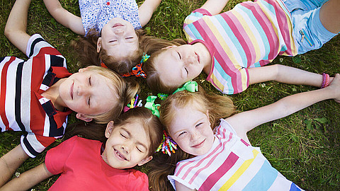 Unsere Projekte Fünf Kinder liegen an einem sonnigen Tag im Kreis auf dem Rasen, die Köpfe aneinander gelehnt, tragen bunte Kleidung und lächeln in die Kamera.