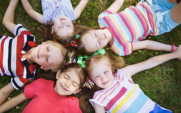 Fünf Kinder liegen an einem sonnigen Tag im Kreis auf dem Rasen, die Köpfe aneinander gelehnt, tragen bunte Kleidung und lächeln in die Kamera.