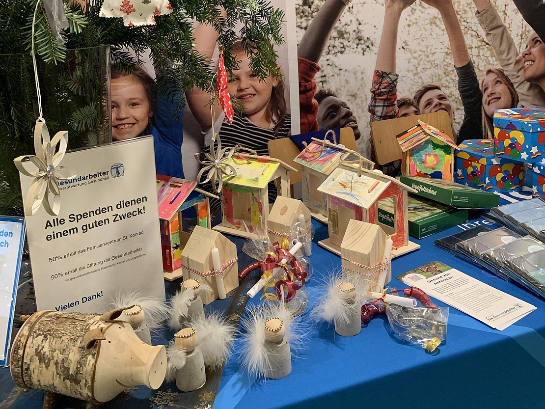 Ein geschmückter Tisch mit handgefertigten Kunstwerken, Weihnachtsschmuck und einer Spendenbox neben einem Weihnachtsbaumzweig. Dahinter wirbt ein Banner mit Kindern, die einen Globus halten, für eine Initiative im Gesundheitsbereich.